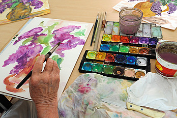 Malkurs Eine ältere Hand hält einen Pinsel über einem gemalten Bild