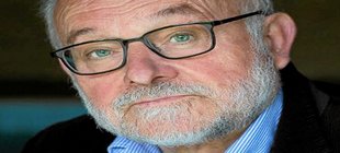 Mann mit blauem Hemd, dunkler Anzugjacke, Brille und Bart vor einem dunklen Hintergrund