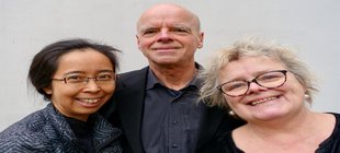 Ein Mann und zwei Frauen in schwarzer Kleidung vor einem grauen Hintergrund