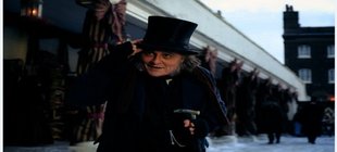 Ebenezer - Eine Weihnachtsgeschichte Person in historischer Kleidung mit Zylinderhut und Gehstock steht in einer verschneiten Straße des 19. Jahrhunderts vor einer Ladenzeile.
