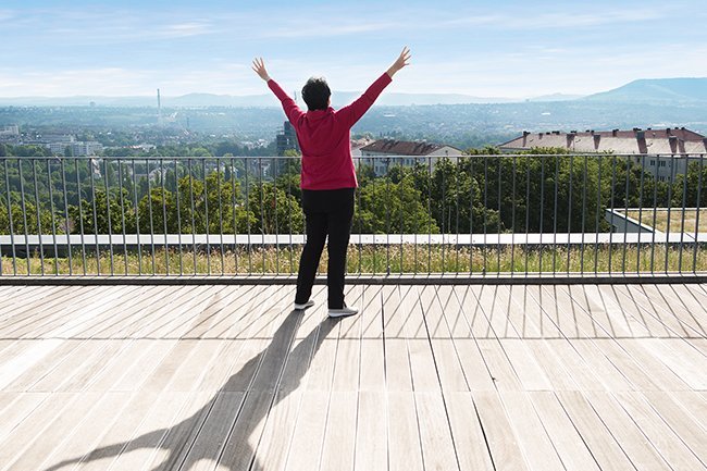 Qi Gong Eine Seniorin steht mit nach oben gestreckten Armen auf einer Terrasse