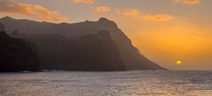 Bergige Küste vor einem farbigen Sonnenuntergang, im Vordergrund das Meer