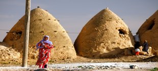 Syrien Vor kegelförmigen Lehmbauten steht eine Frau mit Kopftuch im roten Kleid.