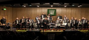 „Mozart, Mancini and more!“ Ein Akkordeon-Orchester mit Dirigent und Schlagzeuger steht auf der beleuchteten Bühne.