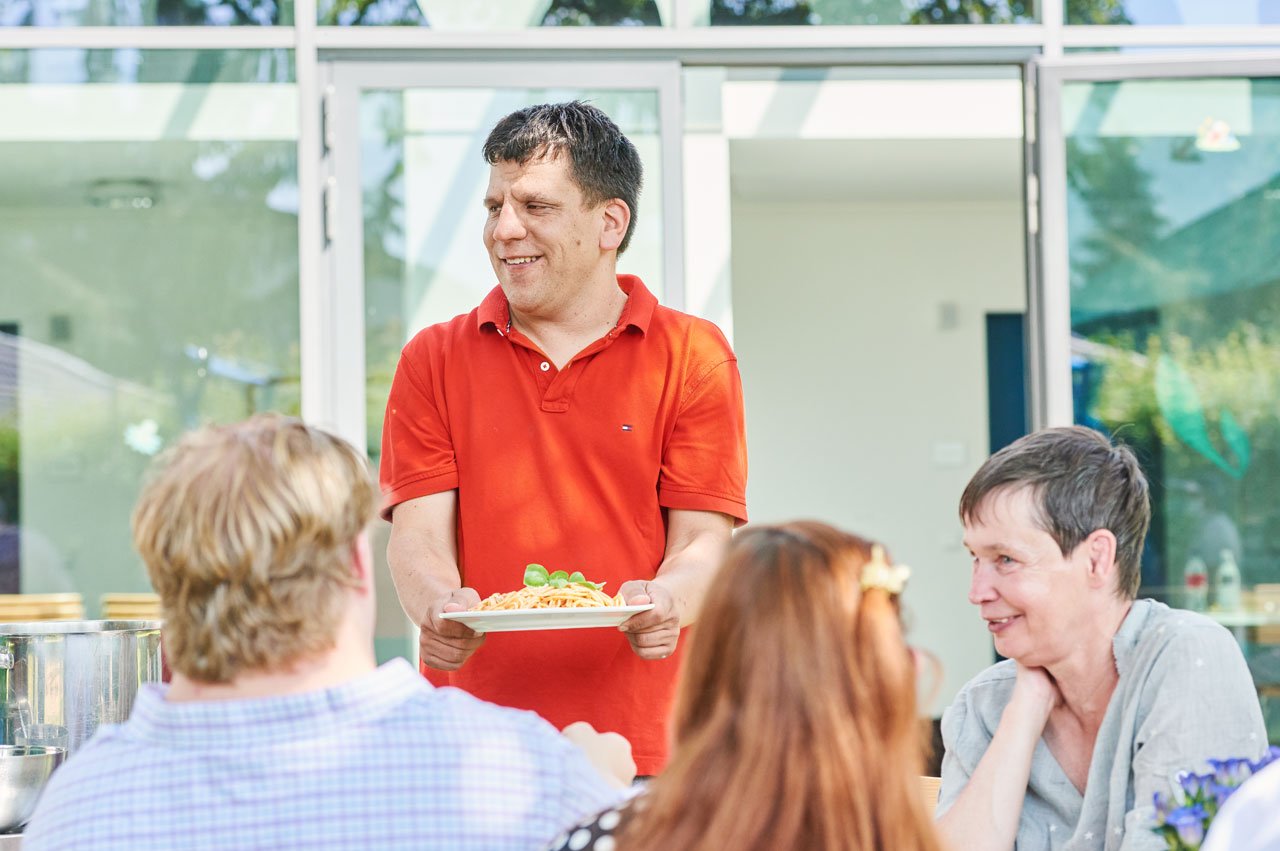 Gemeinschaft Ein Bewohner gesellt sich mit einem Teller Pasta in den Händen zu den anderen Bewohner*innen an einen Tisch im Freien