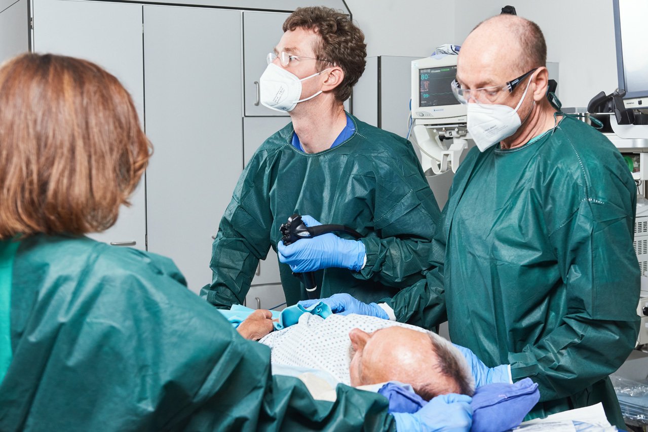Magenspiegelung Ein dreiköpfiges Behandlungsteam steht um einen liegenden Patienten, bei dem eine Magenspiegelung durchgeführt wird.