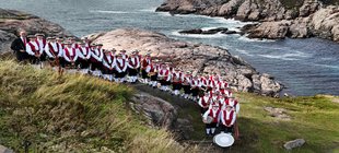 Sommerserenade Große Musikgruppe in roten Westen und historischen Hüten mit Blasinstrumenten auf einem Küstenpfad; im Hintergrund sind Felsen und das Meer zu sehen.