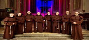 Magic Gregorian Night Acht Personen in braunen Ordensgewändern stehen vor dem Altarraum einer Kirche mit farbig beleuchteten Spitzbogenfenstern und Kreuz im Hintergrund