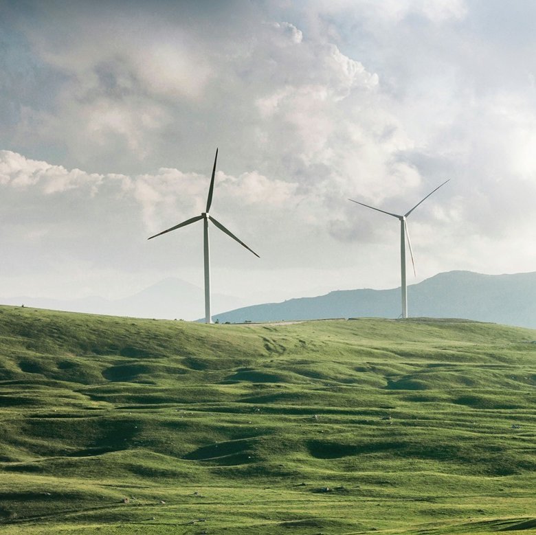 Augustinum Nachhaltigkeit Slider Zwei Windräder vor einer hügeligen Wiesenlandschaft.