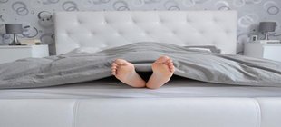 Verteidigung des Mittagsschlafs Zwei Füße lugen unter der grauen Bettdecke eines weißen Bettes hervor