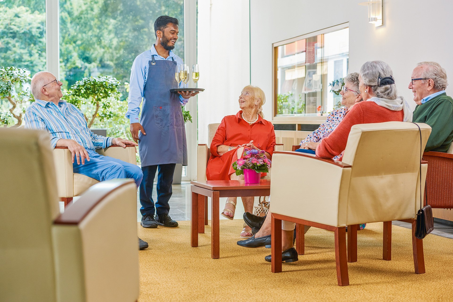 Augustinum Seniorenresidenz Bonn Foyer Eine Gruppen von Senior*innen sitzt gemeinsam im Foyer und ein Mitarbeiter des Augustinum bringt ihnen Sektgläser