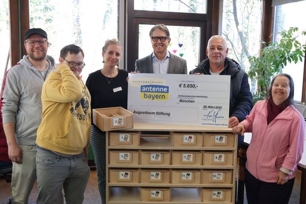 Augustinum Werkstätten Arbeitskisten Ein Gruppenbild mit Beschäftigten und Mitarbeiter*innen der Augustinum Werkstätten, die um die Arbeitskisten und einem Spendenscheck der Antenne-Bayern-Stiftung stehen