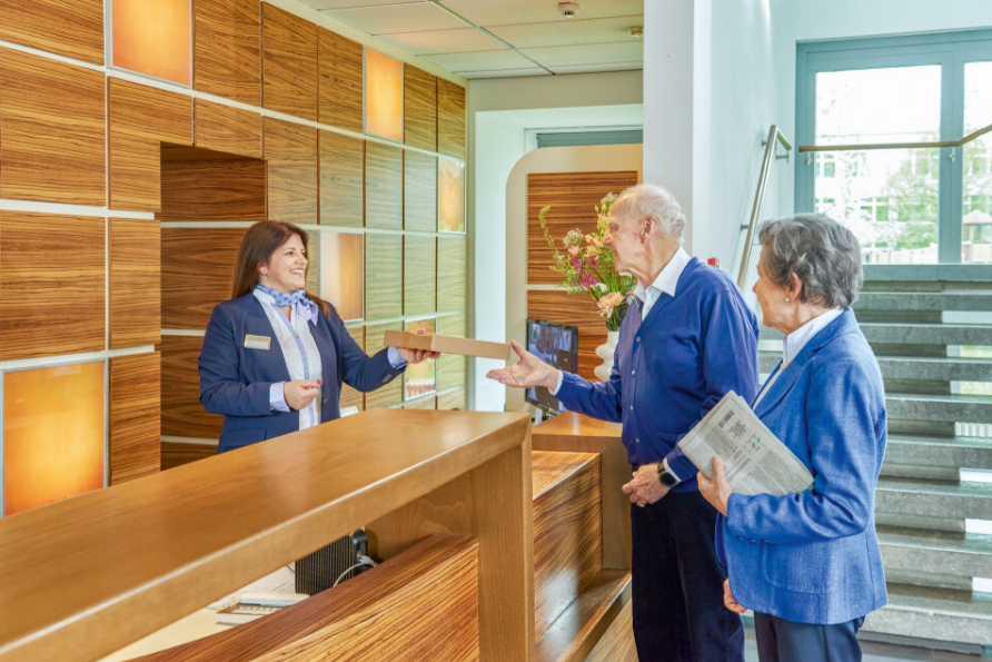 Augustinum Seniorenresidenz Braunschweig Empfang Eine Mitarbeiterin am Empfang des Augustinum übergibt ein Paket an ein Seniorenpaar