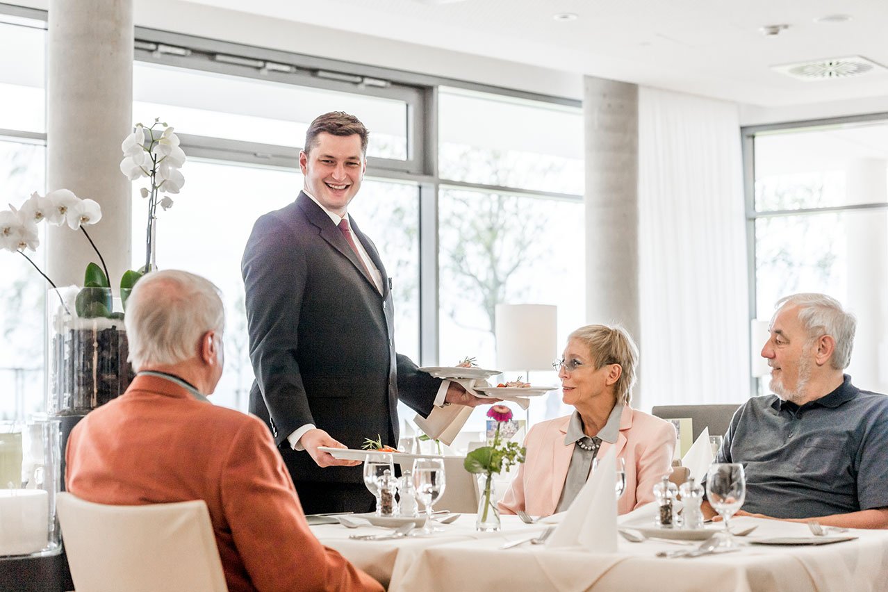 Die Mitarbeiter*innen im Service kennen „ihre“ Gäste und wissen um deren Vorlieben. Kellner serviert Gästen am gedeckten Tisch