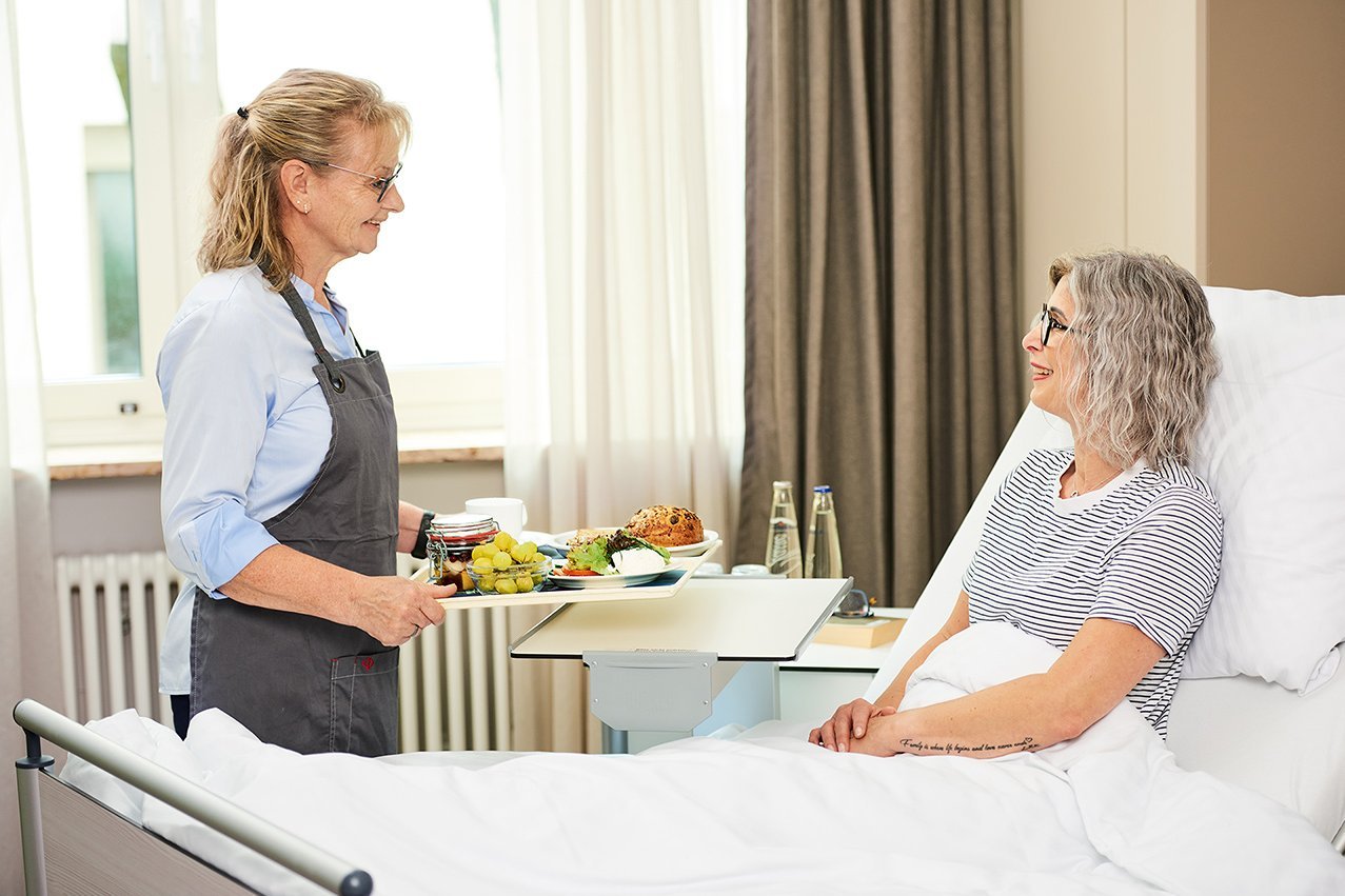 Augustinum Klinik Wahlleistungsstation Mahlzeit Eine Mitarbeiterin der Augustinum Klinik bringt einer im Bett liegenden Patientin ihr Essen