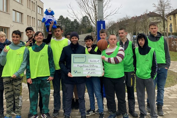 Augustinum Otto-Steiner-Schule Basketballkorb Schüler*innen vor Basketballkorb und Mann von BBBank, der Spendenscheck in den Händen hält, stehen vor einem Basketballkorb im Pausenhof