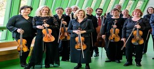 Ein Abend voller Operndramatik und Streicherglanz Die Musiker*innen des Kammerorchesters Arcata stehen mit ihren Instrumenten in der Hand in schwarzer Konzertkleidung vor einer Fensterfront.
