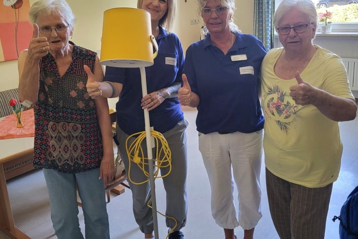 Augustinum Fördermagazin Tovertafel Ein Bild einer Mitarbeiterin des Augustinum die eine Tovertafel hält, und drei Seniorinnen, die einen Daumen nach oben zeigen