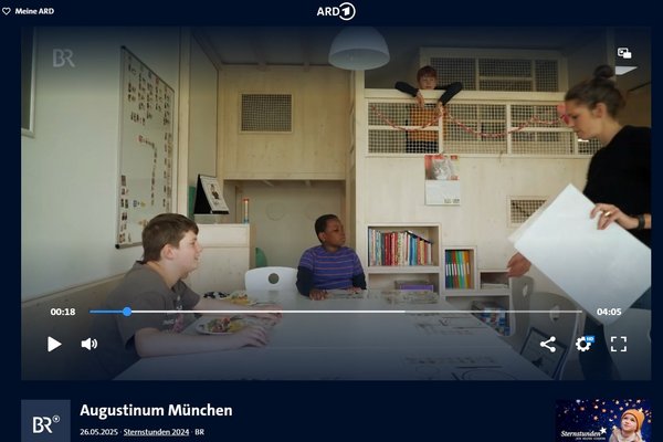 Augustinum Tagesstätten im Fernsehen Ein Raum mit Holzverkleidung und 2 Ebenen ist zu sehen. An einem Tisch sitzen zwei Kinder. Rechts steht eine Frau mit großen Papierblättern in der Hand