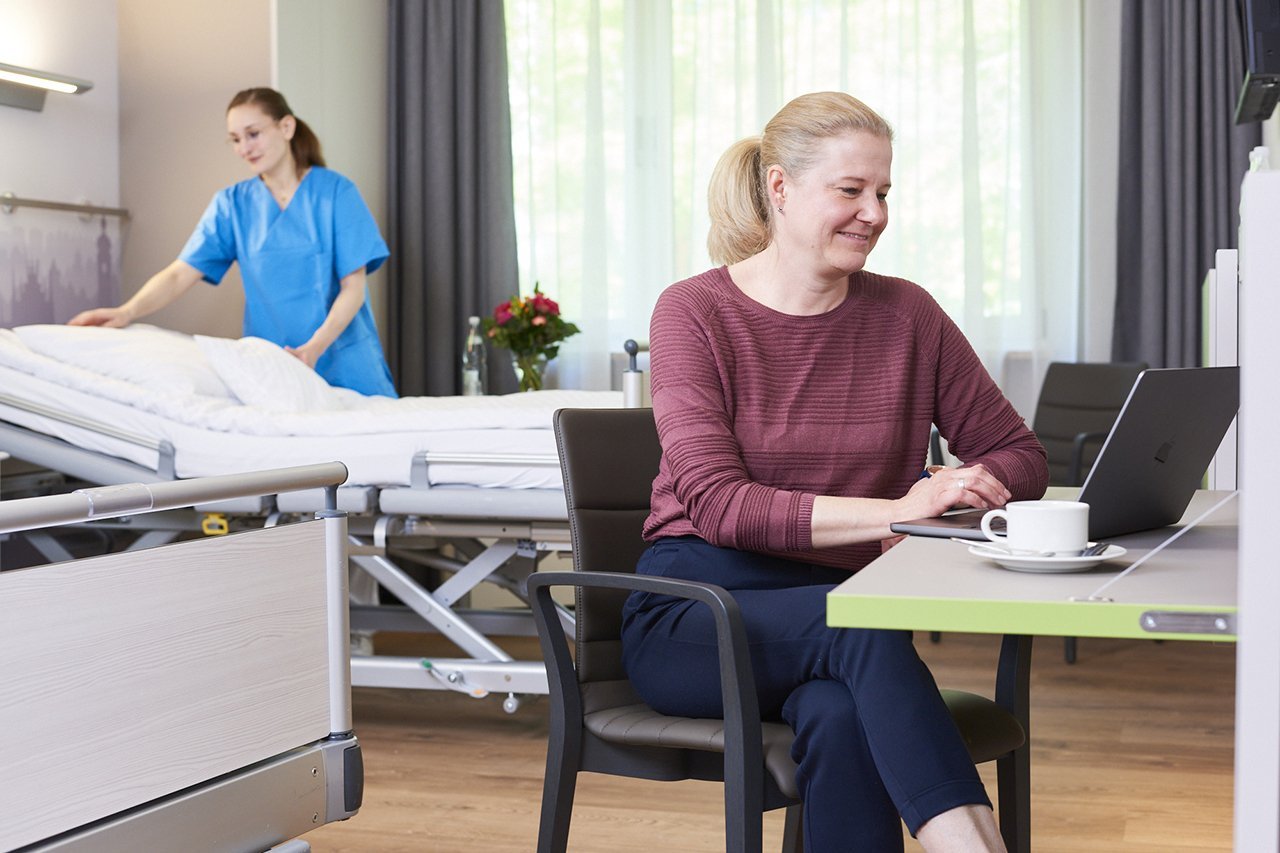 Augustinum Klinik Wahlleistungsstation Zimmer Eine Frau sitzt an einem Tisch vor ihrem Laptop in einem Zimmer der Wahlleitungsstation und im Hintergrund macht eine Mitarbeiterin der Augustinum Klinik das Bett