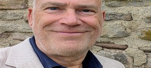 Ein Mann in dunklem Hemd und hellem Anzug vor einer Steinmauer