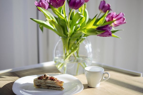 Ausstattung der Patientenzimmer Auf einem Tisch steht ein Strauß violetter Tulpen neben einem Stück Kuchen und einer Tasse Kaffee