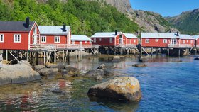 Rot gestrichene Holzhäuser auf den Lofoten; im Vordergrund Wasser, im Hintergrund bewaldete Berge