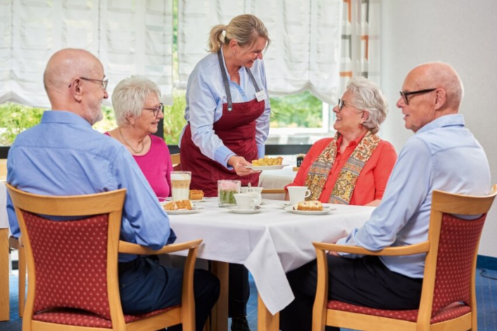 Augustinum Seniorenresidenz Dortmund Restaurant Vier Senior*innen sitzen an einem Tisch im Restaurant und eine Mitarbeiterin serviert ihnen Kuchen