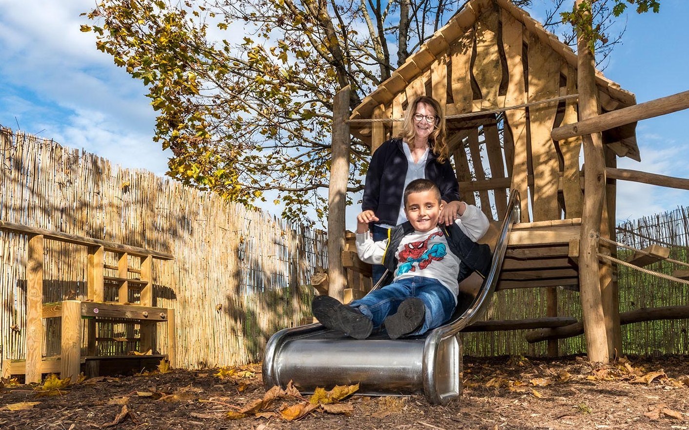 fröhlicher Junge auf einer Rutsche, im Hintergrund eine Erzieherin