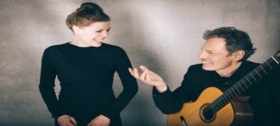 Ein Mann mit Gitarre redet mit einer Frau, beide schwarz gekleidet, vor einer grauen Wannd