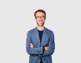 Mann mit Brille in blauem Blazer und schwarzem Shirt vor einem grauen Hintergrund
