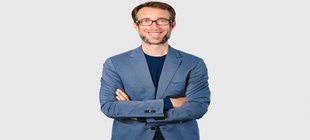 Mann mit Brille in blauem Blazer und schwarzem Shirt vor einem grauen Hintergrund