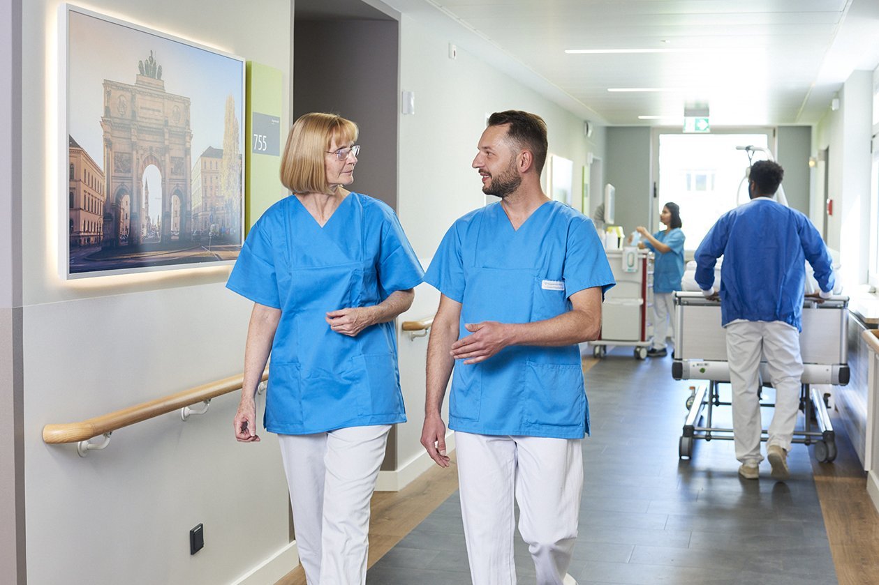 Augustinum Klinik Wahlleistungsstation Flur Zwei Mitarbeiter*innen der Augustinum Klinik unterhalten sich im Flur einer Wahlleistungsstation, während im Hintergrund weitere Mitarbeiter*innen bei der Arbeit zu sehen sind
