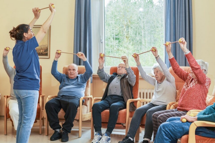 Augustinum Berchtesgadener Land Tagespflege Gemeinschaftsaktivität Eine Mitarbeiterin der Tagespflege zeigt einer Gruppe von Senior*innen eine Übung