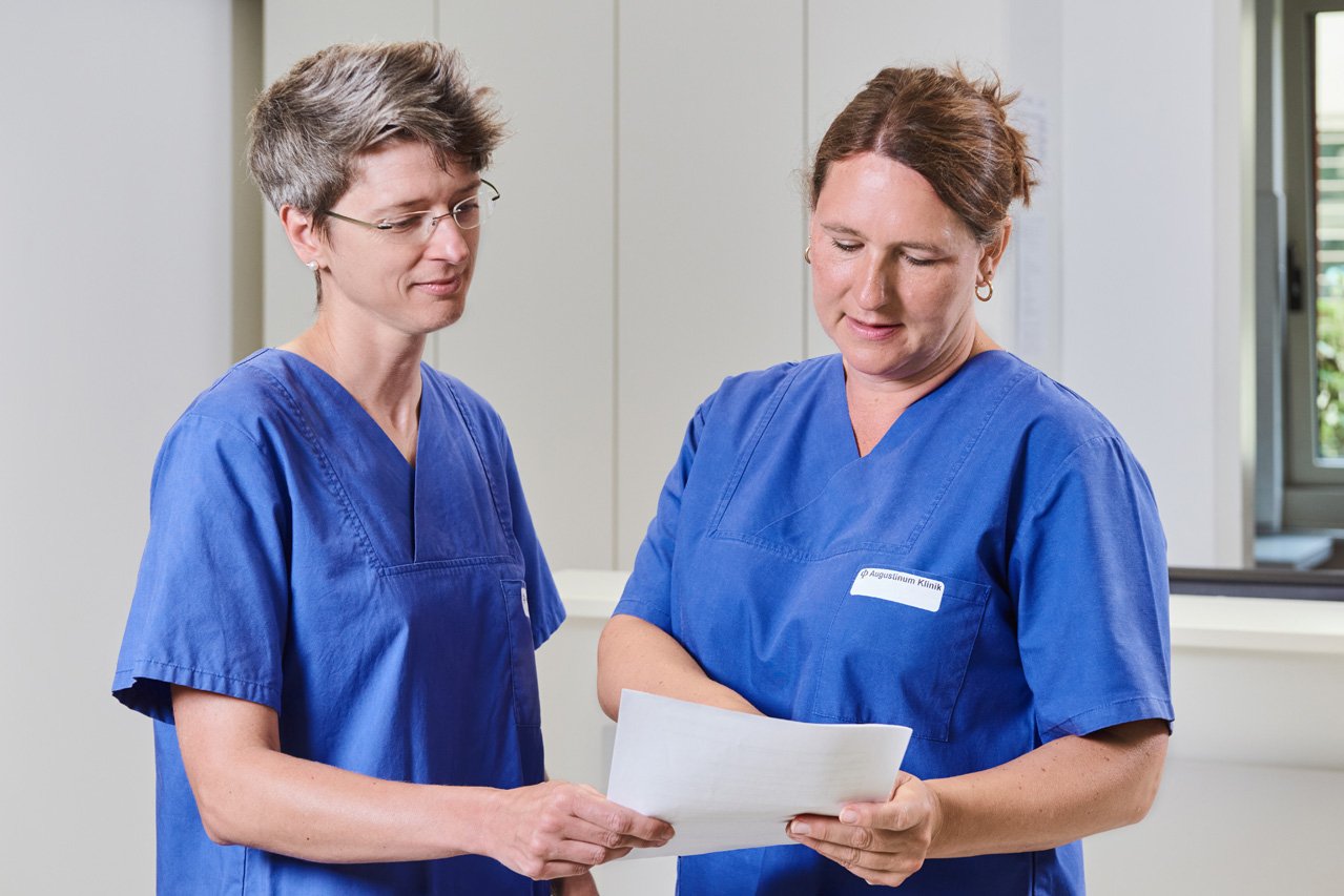 Besprechung Ärztinnen Zwei Ärztinnen stehen nebeneinander und sehen gemeinsam auf ein Schriftstück.