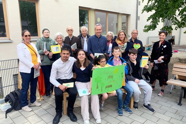 Einweihung mit Parkbank-Paten 5 Kinder und Jugendliche sitzen auf einer Parkbank und halten ein Danke-Schild in die Kamera. Im Hintergrund stehen 10 Erwachsene, die Bänke gespendet haben