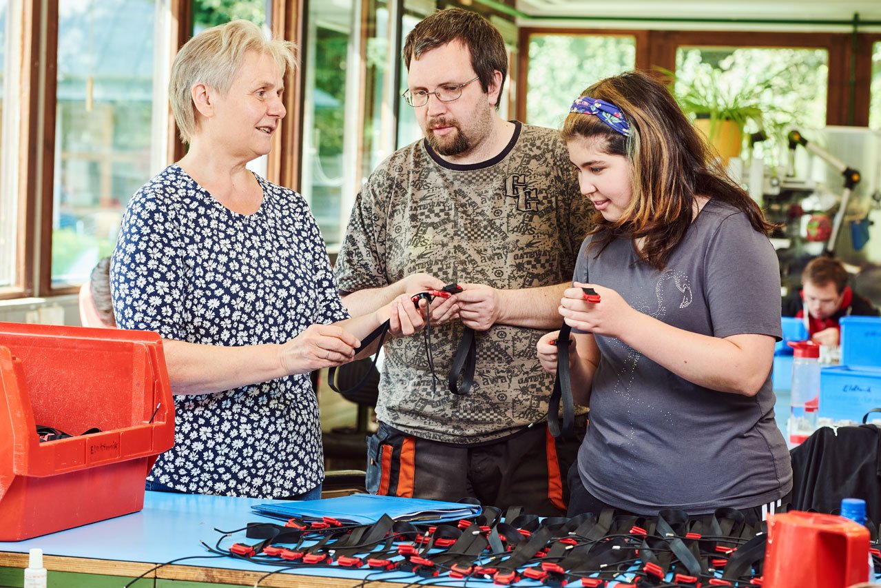 Klient*innen der Elektronikabteilung Eine Mitarbeiterin erklärt zwei Klient*innen einen Arbeitsauftrag in der Elektronikabteilung