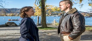Stiller Ein Mann und eine Frau stehen sich an einem Seeufer gegenüber. Im Hintergrund sind Bäume mit herbstlichem Laub, mehrere Vögel über dem Wasser sowie ein kleines Boot auf dem See zu sehen.