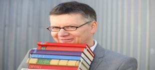 Mann mit Brille und einem Bücherstapel vor einer grauen Wand